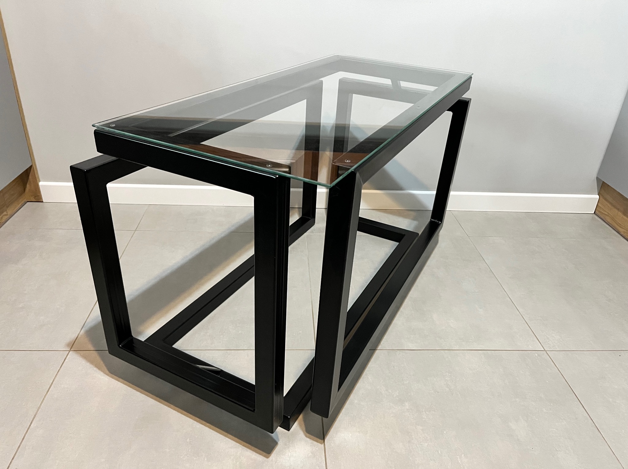 Nowoczesny stolik kawowy z czarną, metalową ramą i szklanym blatem, ustawiony na jasnych płytkach podłogowych. Minimalistyczny design, widoczne detale łączeń.