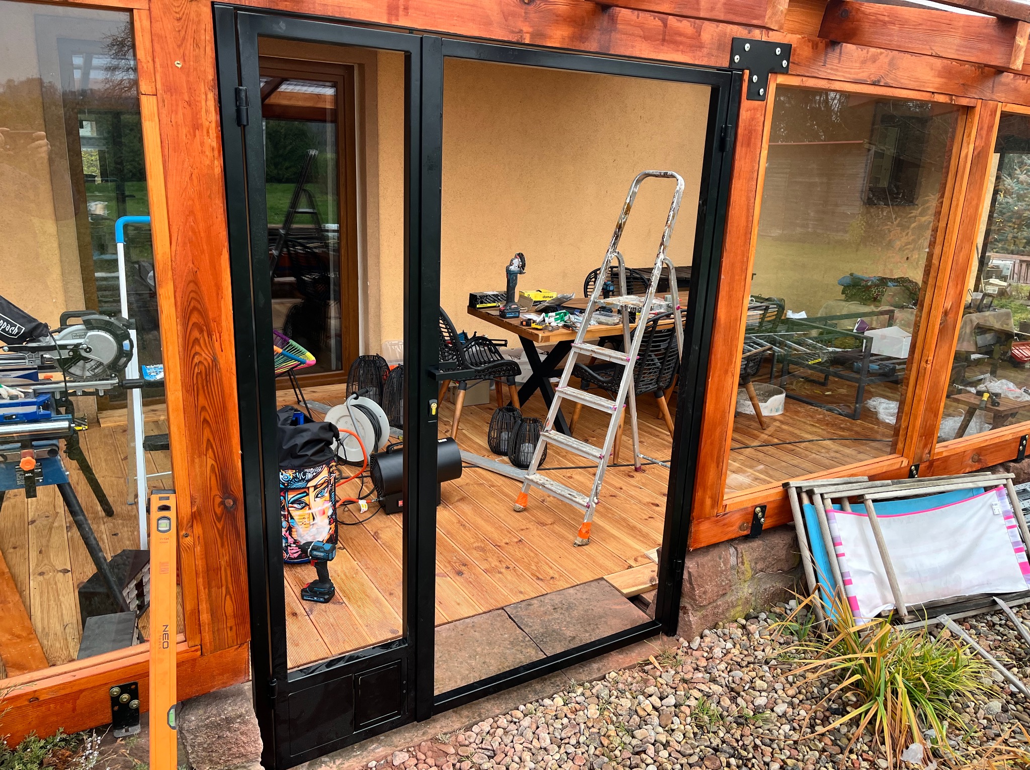 Częściowo zamontowana, czarna, metalowa konstrukcja w drewnianej ramie, prawdopodobnie szklarni. W tle narzędzia i drabina na drewnianej podłodze.