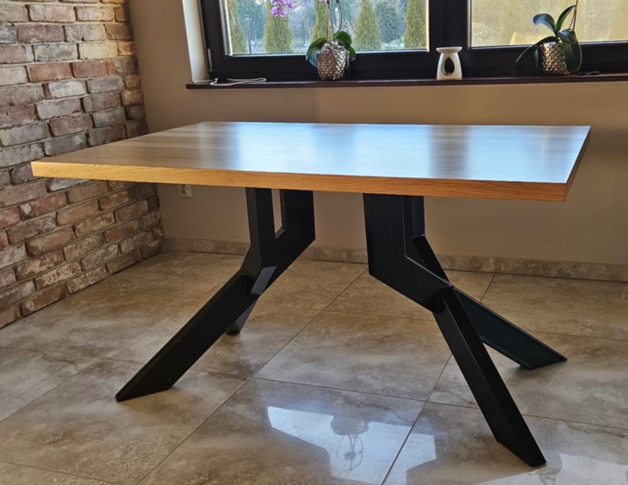 Stół z blatem z jasnego drewna i czarnymi, metalowymi nogami o geometrycznym kształcie. W tle ceglana ściana i okno z roślinami doniczkowymi.