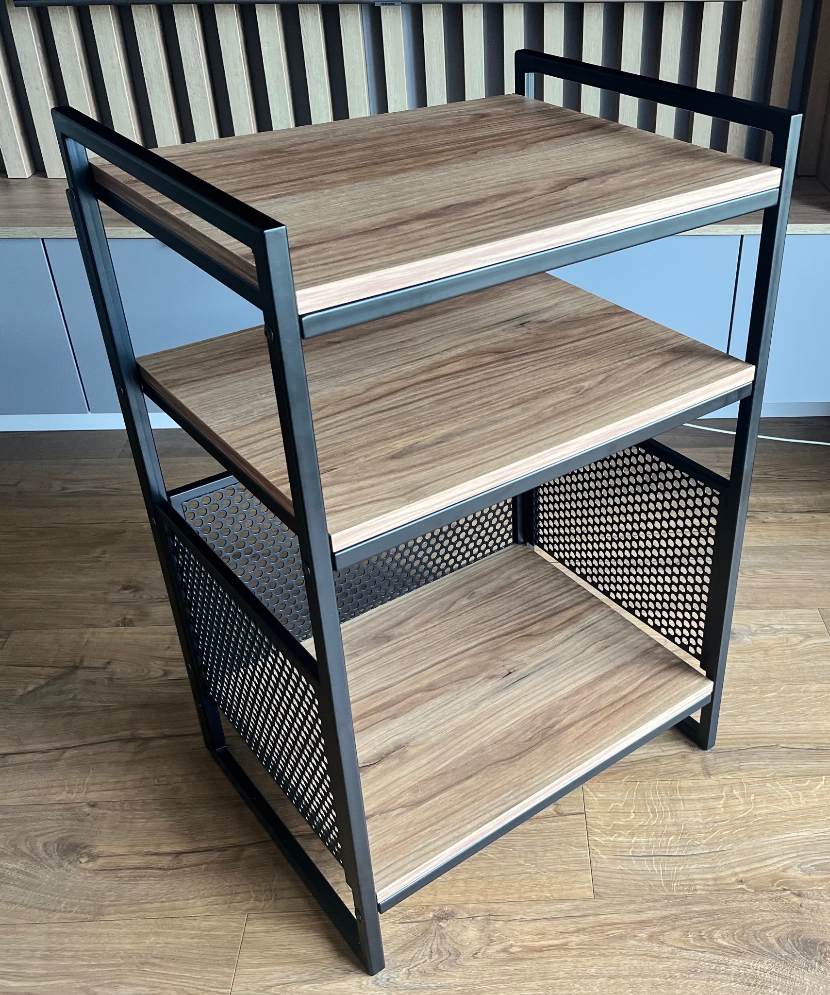 Trzypoziomowy regał loftowy z drewnianymi półkami i metalową siatką po bokach, stojący na drewnianej podłodze. Minimalistyczny design.