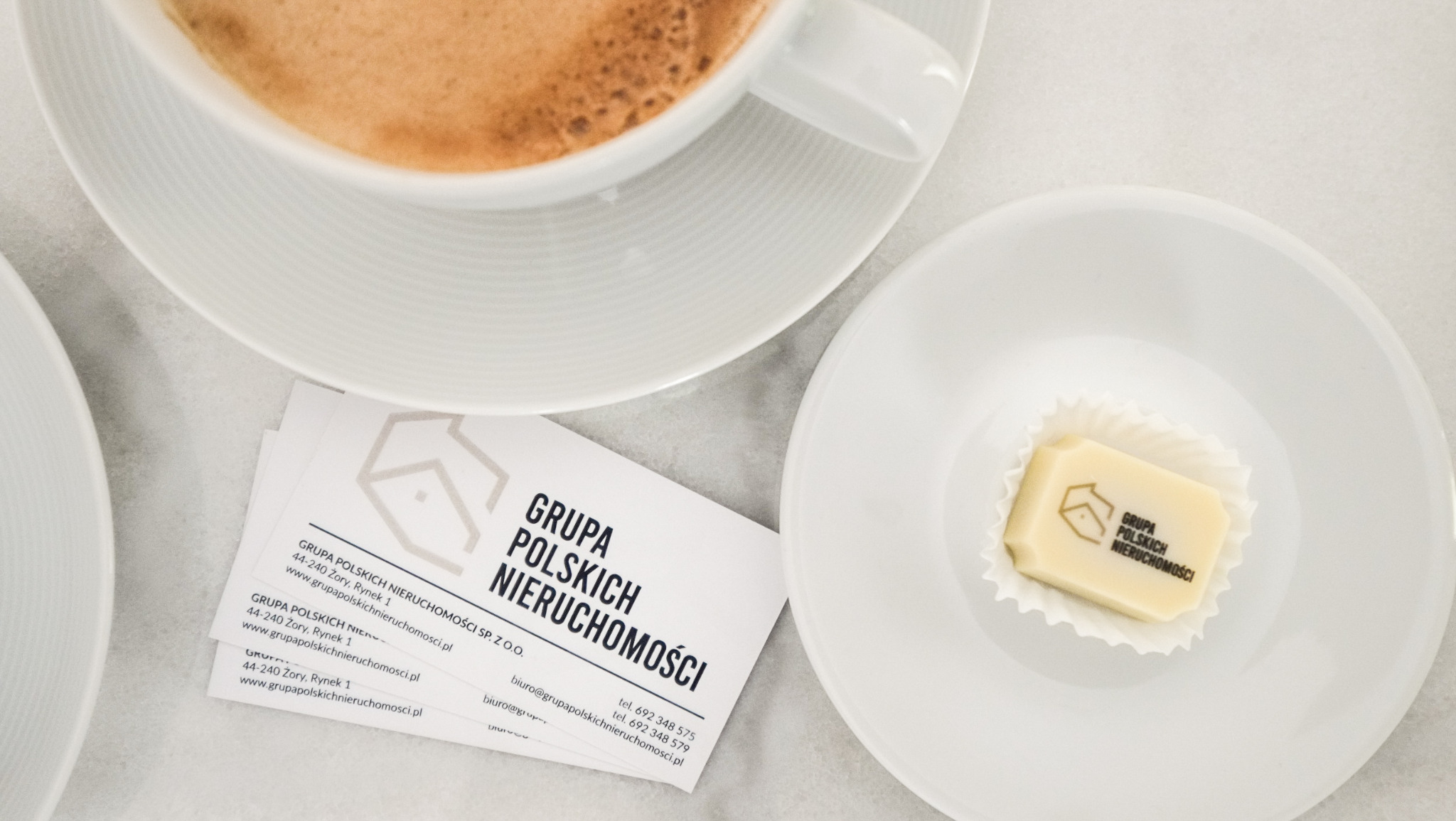 Kawa, wizytówki i czekoladka z logo Grupy Polskich Nieruchomości na białym tle. Elegancki branding i zaproszenie do współpracy.