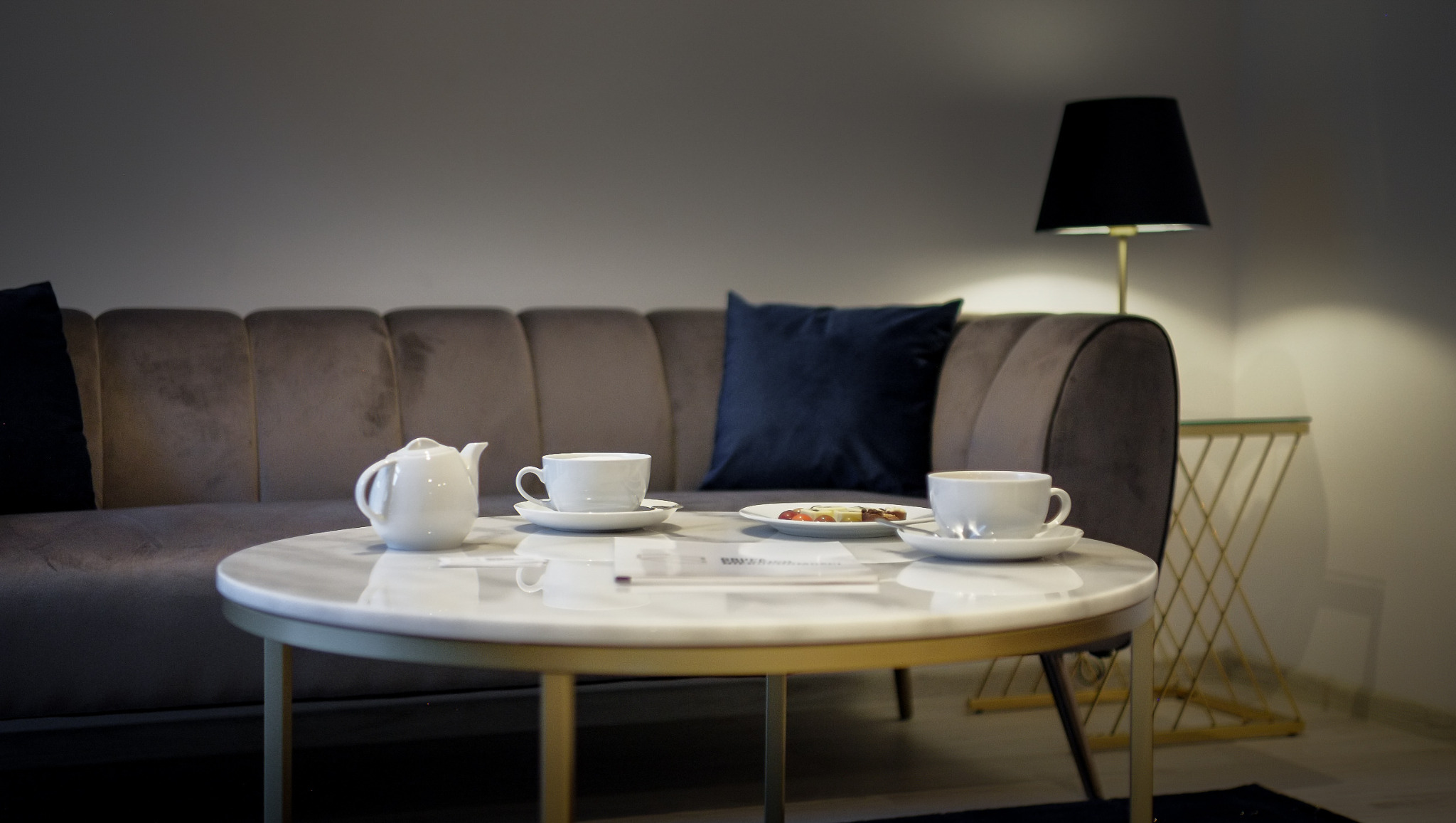 Stylowy salon z sofą, stolikiem kawowym z marmurowym blatem, serwisem do herbaty i lampą. Przytulne wnętrze, idealne do relaksu i spotkań.