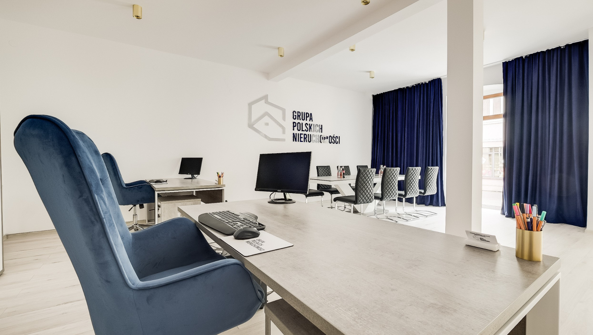 Jasne biuro agencji nieruchomości z logo na ścianie, biurkami, krzesłami i zasłonami w odcieniach granatu. Nowoczesne wnętrze z elementami wyposażenia.