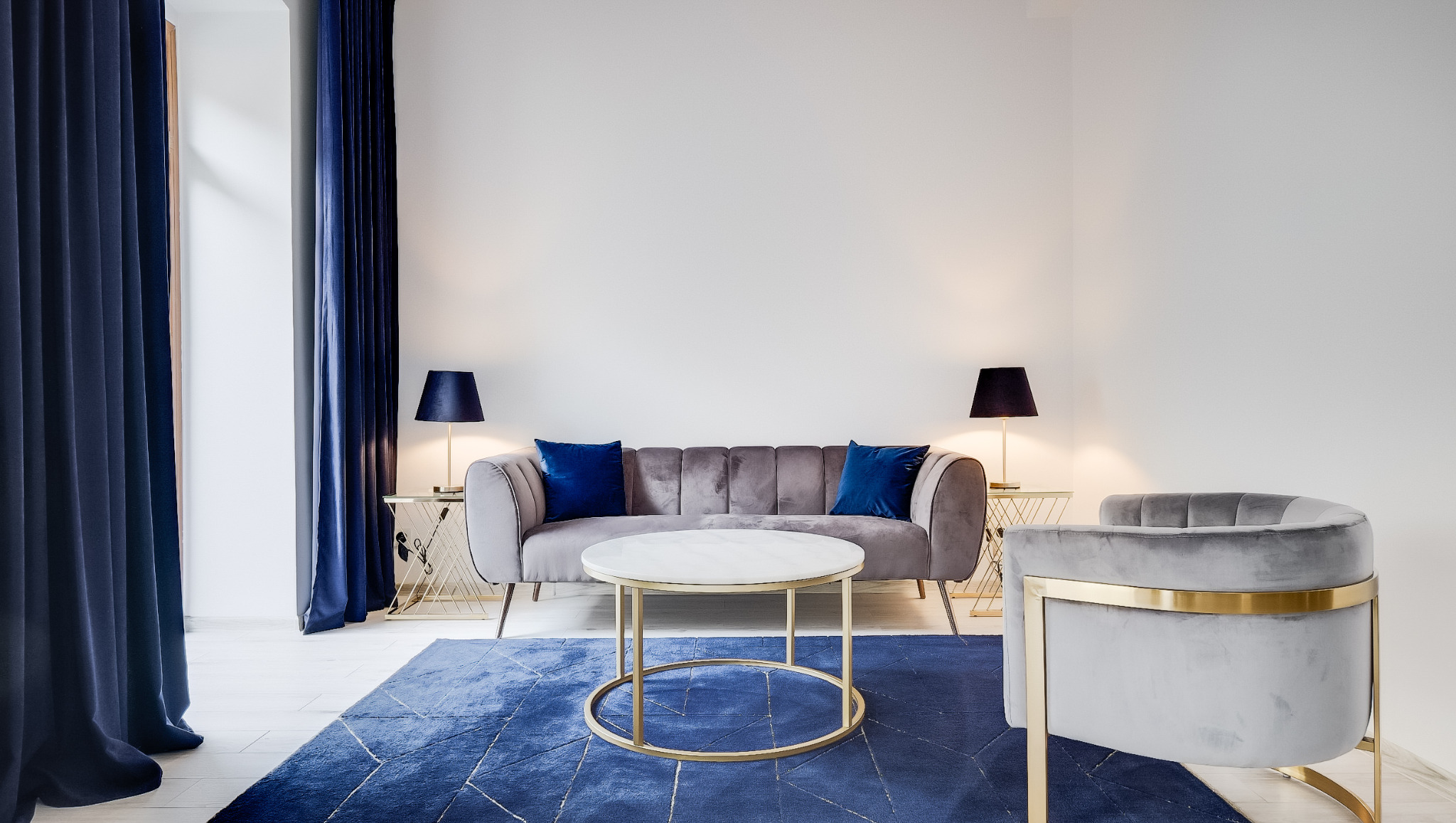 Stylowe wnętrze: szara sofa z niebieskimi poduszkami, marmurowy stolik kawowy na złotym stelażu, niebieski dywan i zasłony. Nowoczesny design z lampkami nocnymi.