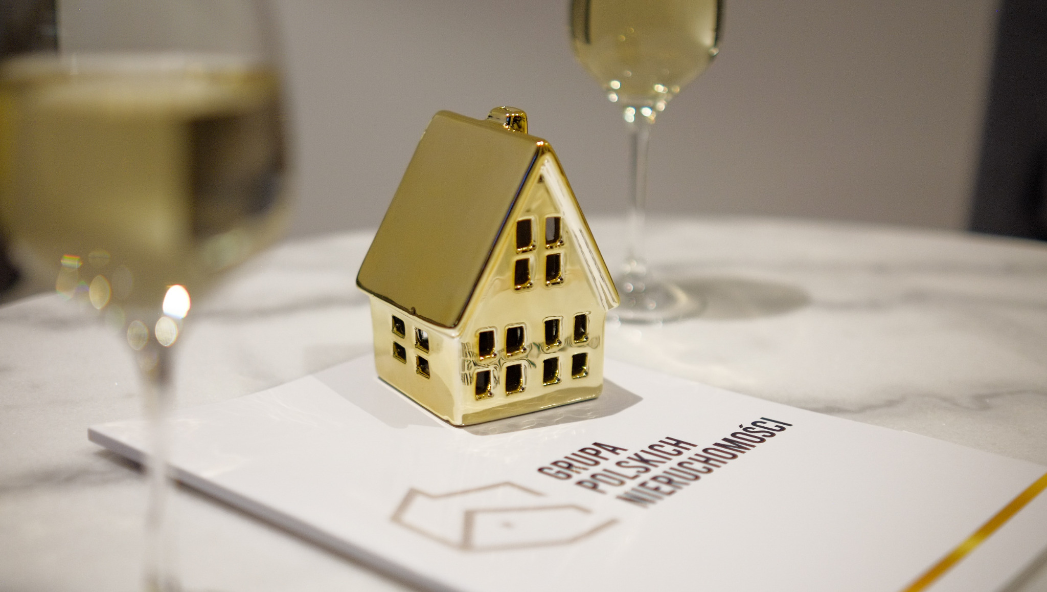 Złota figurka domu na ulotce 'Grupa Polskich Nieruchomości' z kieliszkami szampana w tle. Elegancka prezentacja oferty nieruchomości.