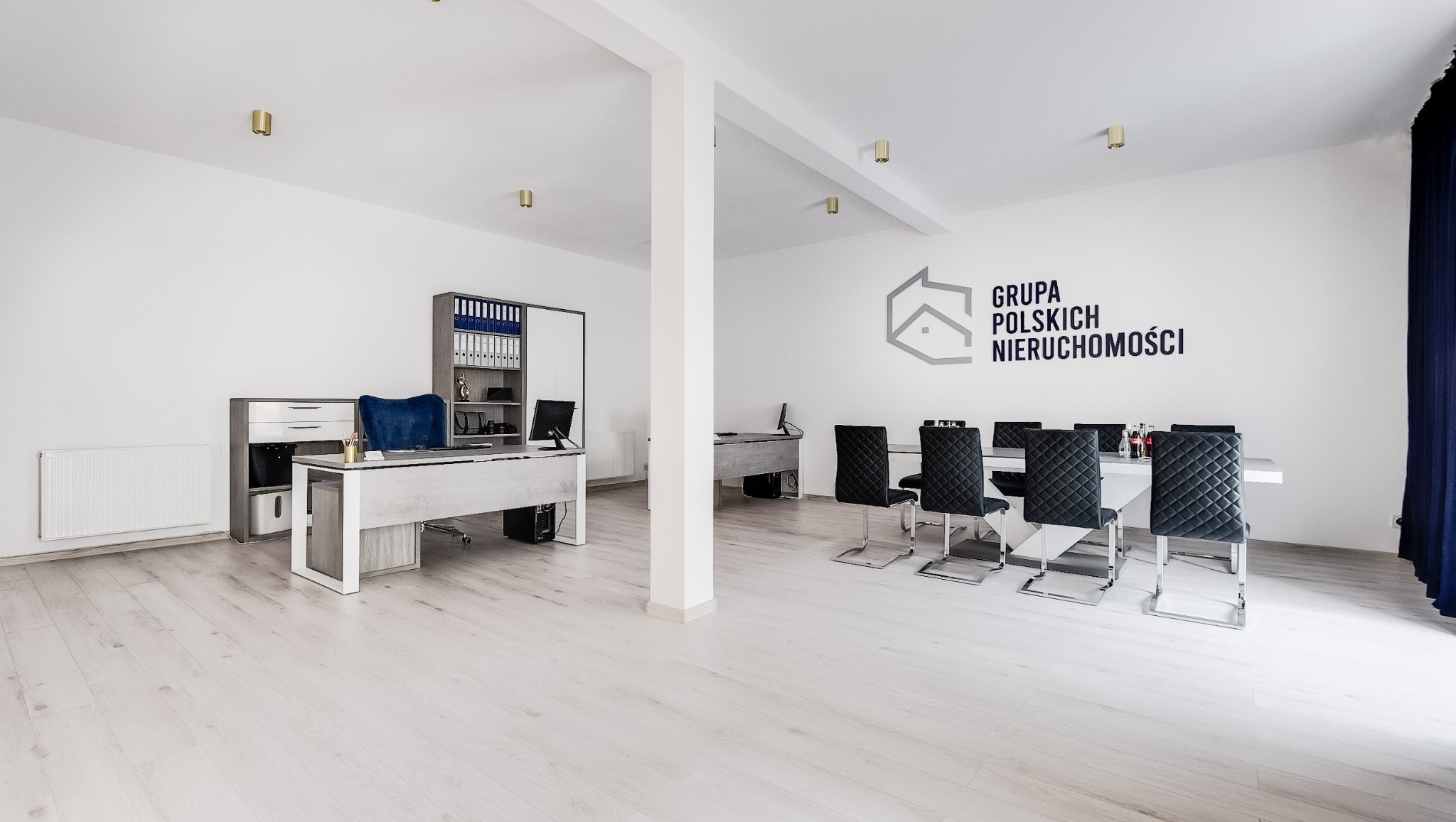 Jasne biuro agencji nieruchomości z logo 'Grupa Polskich Nieruchomości', biurkami, krzesłami i regałami. Minimalistyczny design, białe ściany i podłogi.