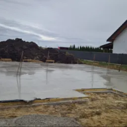 Wylany fundament pod budynek jednorodzinny, widoczne wystające pręty zbrojeniowe i drewniane szalunki. W tle sterta ziemi i budynek w stanie surowym.