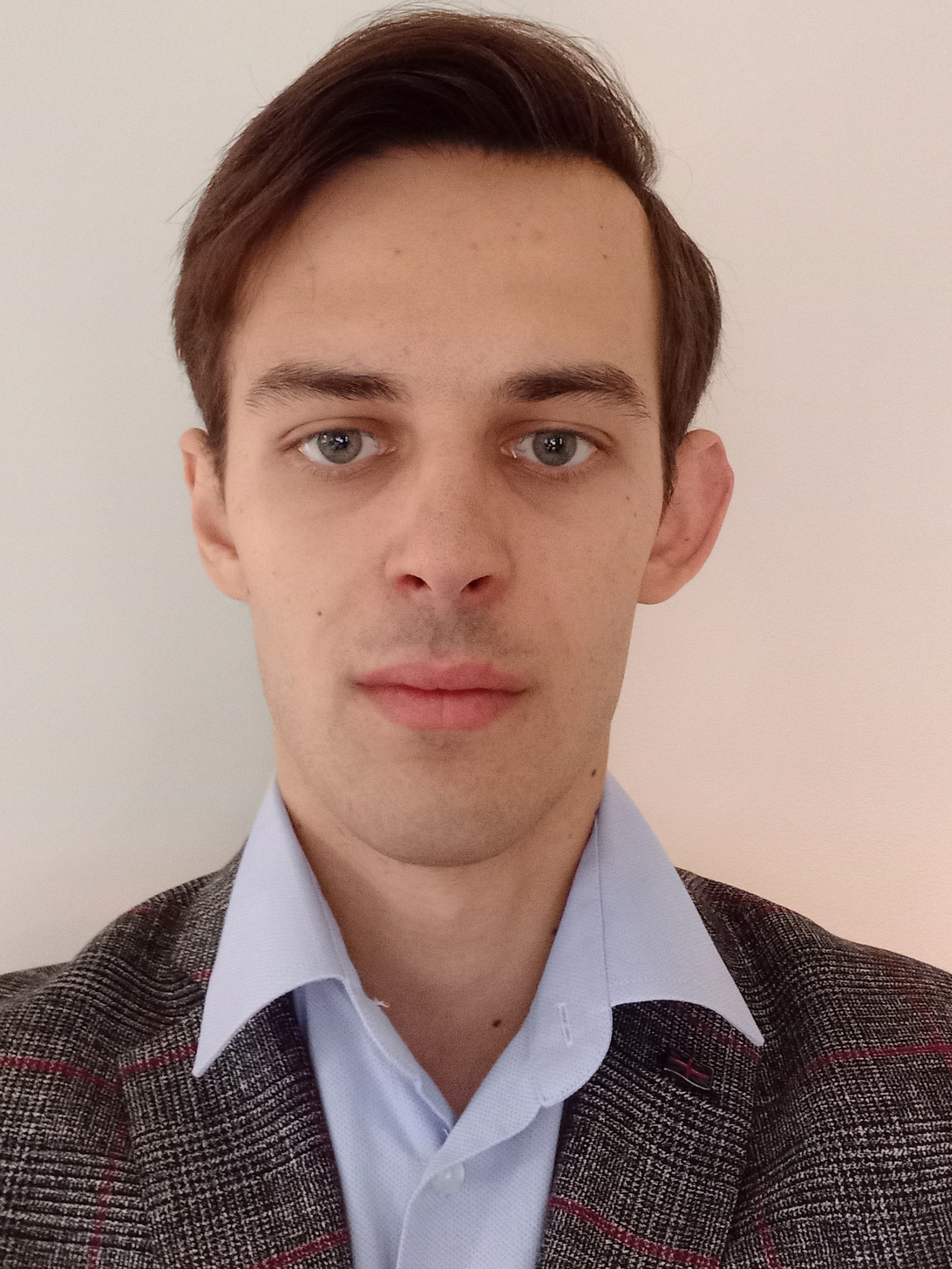 Portret młodego mężczyzny w tweedowej marynarce i błękitnej koszuli, z delikatnym uśmiechem, na jasnym tle. Widoczne detale tkaniny i subtelne oświetlenie.