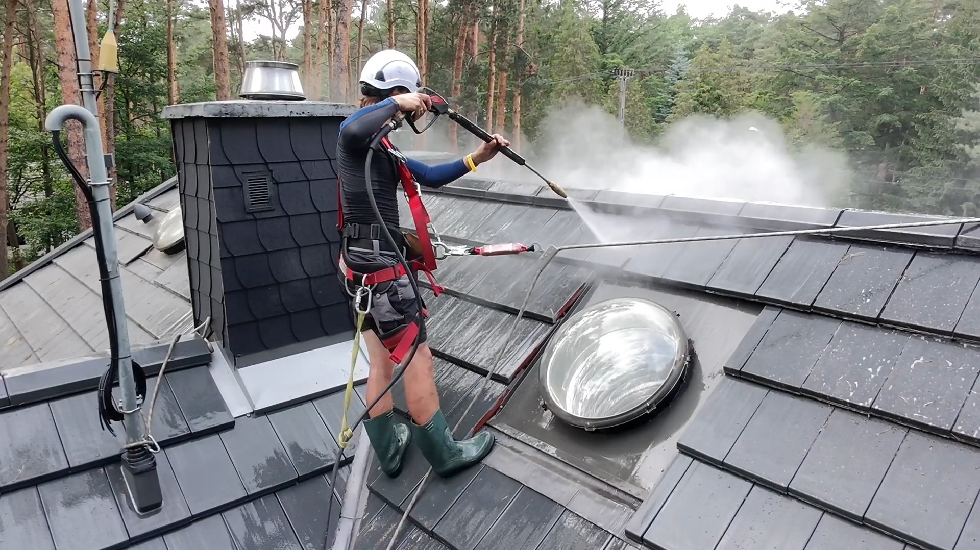 Pracownik w kasku i uprzęży, z bliska myje ciemny dach z dachówki za pomocą myjki ciśnieniowej. Widoczne drzewa w tle i okno dachowe.