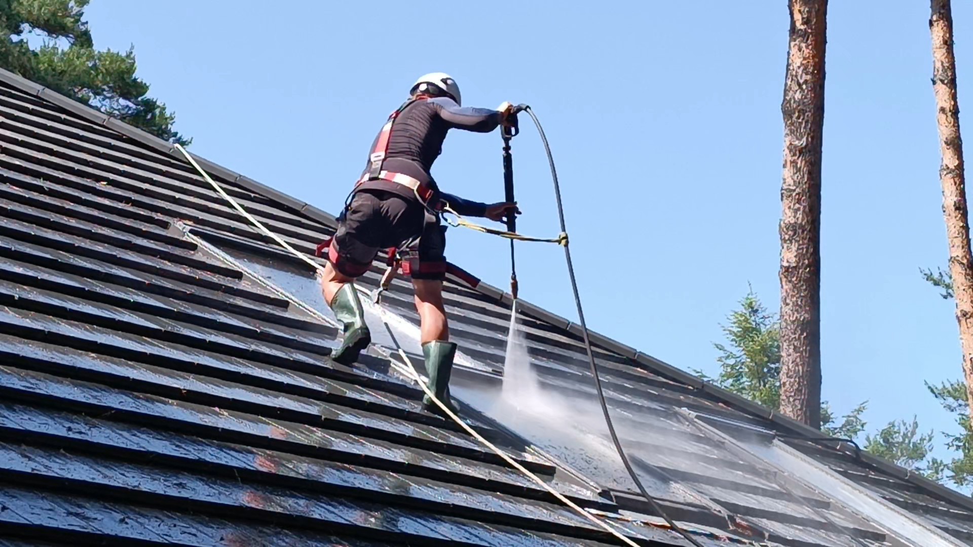 Pracownik w uprzęży, z bliska, czyści ciemny dach z użyciem myjki ciśnieniowej. W tle błękitne niebo i fragmenty drzew. Widoczne strumienie wody.