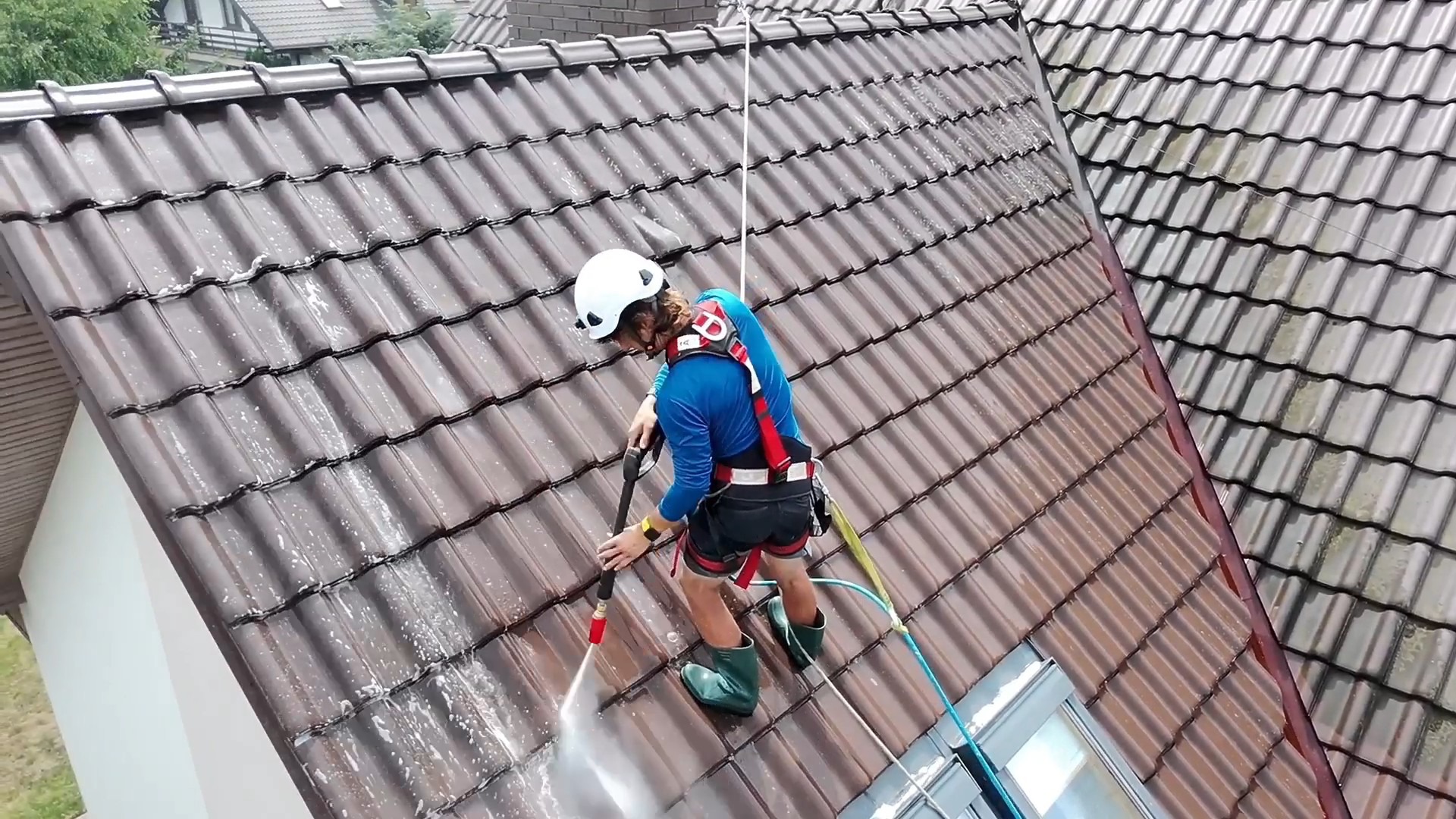 Pracownik w kasku i uprzęży, z bliska czyści dach z brązowej dachówki za pomocą myjki ciśnieniowej. Widoczne zabrudzenia i strumień wody. Ujęcie z góry.