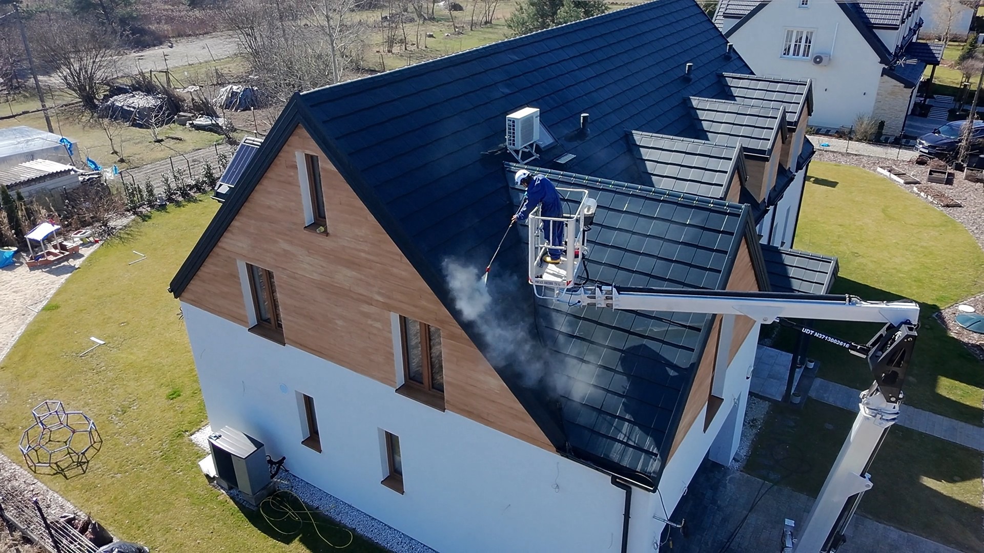 Pracownik w podnośniku czyści ciemny dach domu jednorodzinnego za pomocą myjki ciśnieniowej. Widoczny strumień wody i para. Dachówka ceramiczna. Ujęcie z góry.