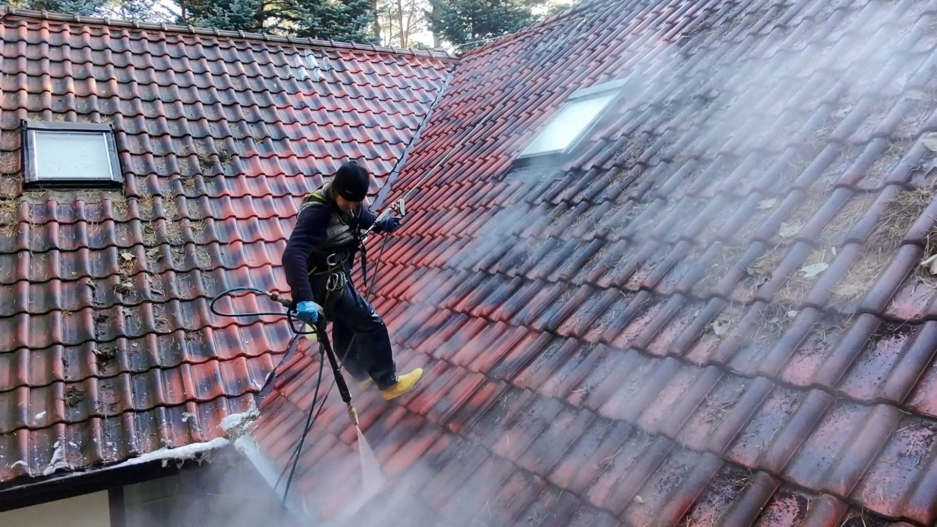 Pracownik w uprzęży czyści dachówkę z zanieczyszczeń za pomocą myjki ciśnieniowej. Widoczny efekt czyszczenia - część dachu umyta, część jeszcze zabrudzona.