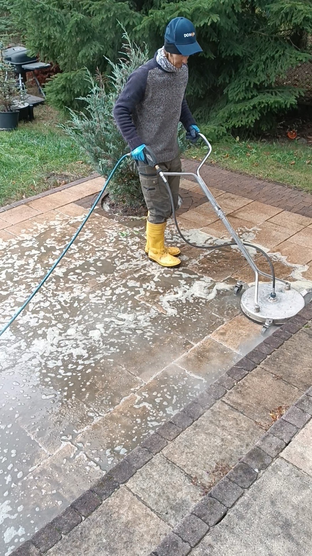 Pracownik w żółtych gumowcach czyści kostkę brukową za pomocą myjki ciśnieniowej. Widoczne zabrudzenia i piana na powierzchni kostki. W tle zieleń ogrodu.