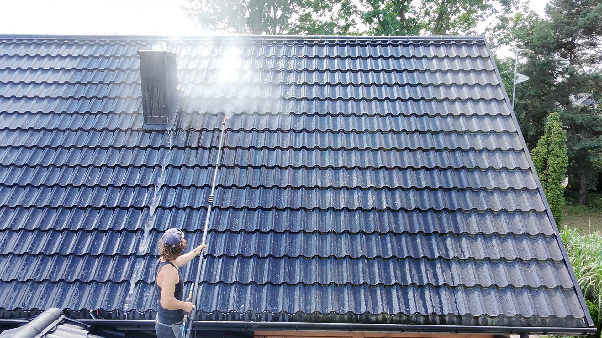 Częściowo umyty, ciemny dach z dachówki ceramicznej, z widocznym kominem. Osoba w trakcie pracy z lancą ciśnieniową. Widoczne efekty czyszczenia.