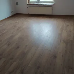 Ukończona podłoga z paneli w jasnym, pustym pokoju z oknem i grzejnikiem. Widoczne listwy przypodłogowe i gładkie ściany. Naturalne światło podkreśla wz&oacute;r drewna.