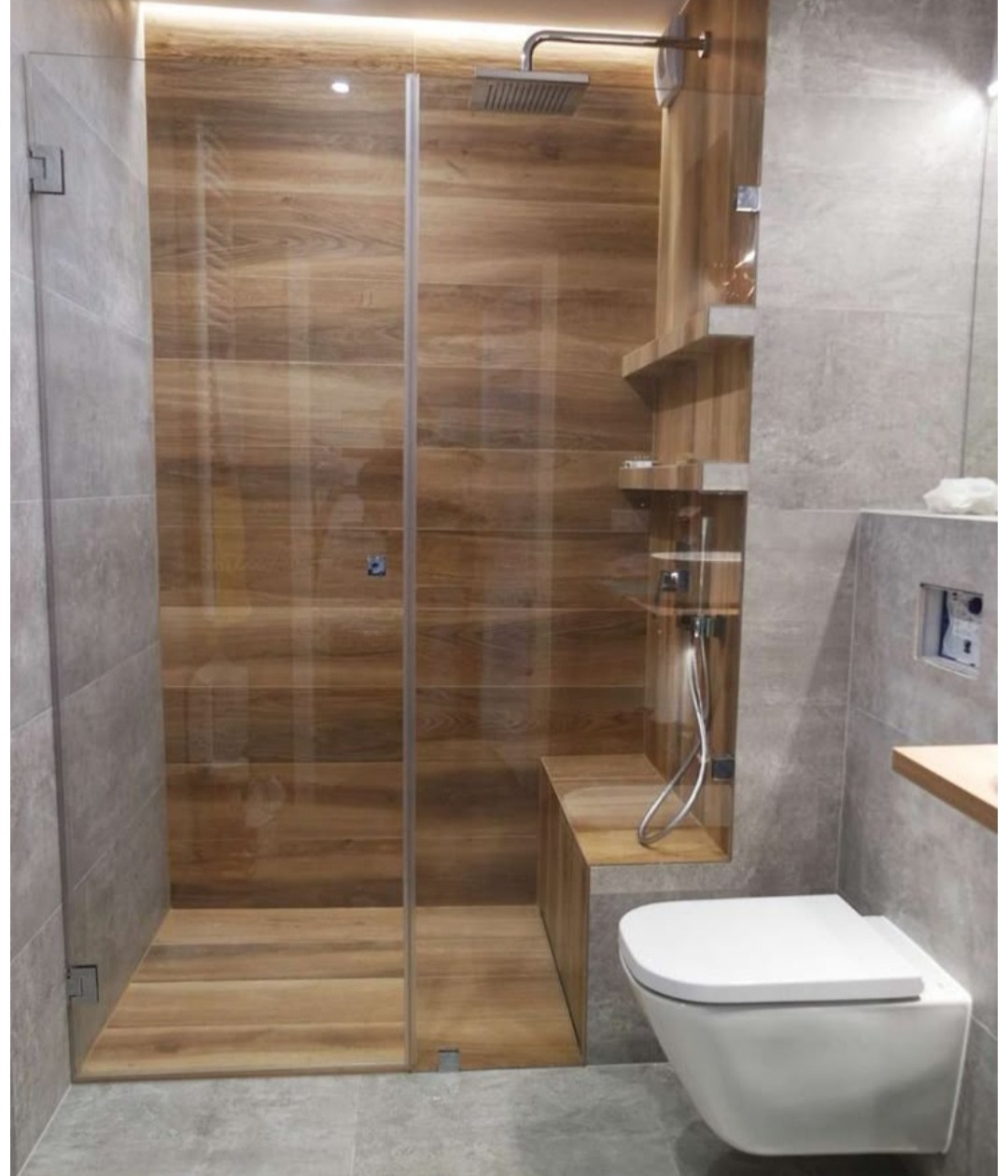Nowoczesna kabina prysznicowa walk-in z drewnopodobnymi płytkami, szklaną ścianką i półkami wnękowymi. Uwagę zwraca minimalistyczna toaleta podwieszana.