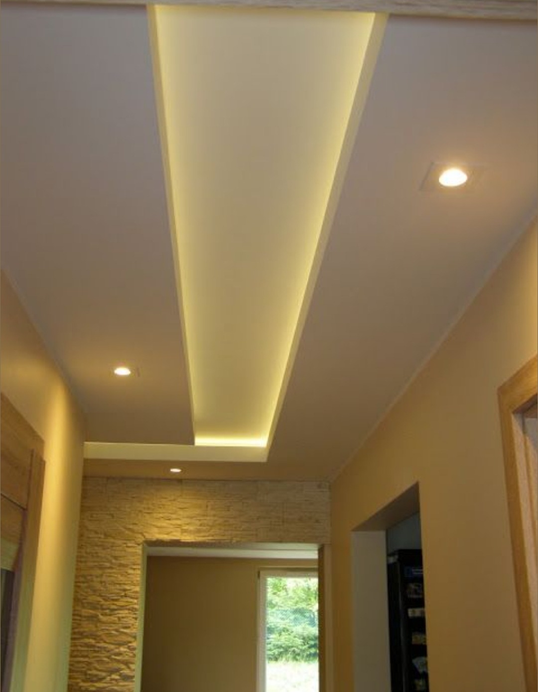 Podwieszany sufit z oświetleniem LED w wąskim korytarzu, wykończony kamieniem dekoracyjnym nad przejściem. Widoczne punktowe światła i beżowe ściany.