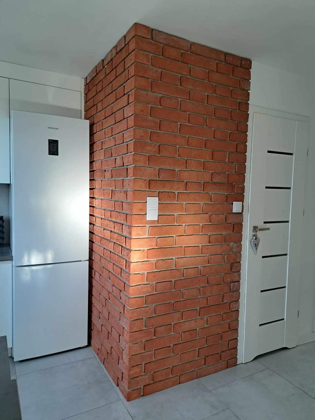 Ceglana ściana dekoracyjna w jasnym wnętrzu, widoczna obok białej lodówki i drzwi. Element wykończenia wnętrza z użyciem cegły.