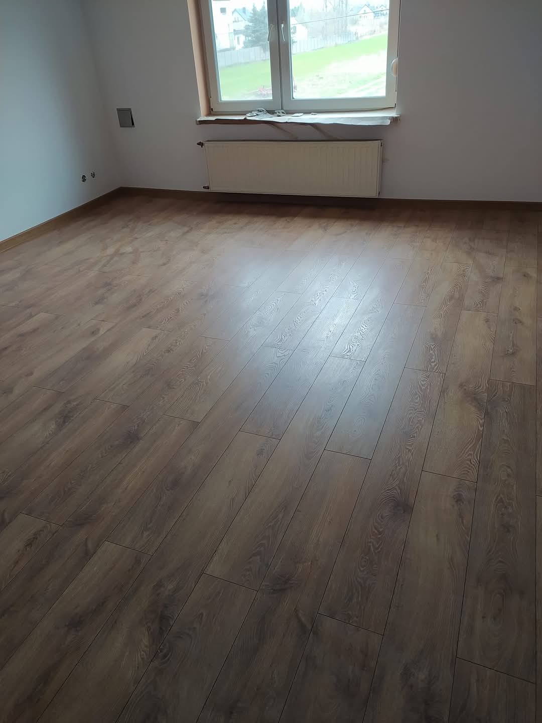 Ukończona podłoga z paneli w jasnym, pustym pokoju z oknem i grzejnikiem. Widoczne listwy przypodłogowe i gładkie ściany. Naturalne światło podkreśla wzór drewna.