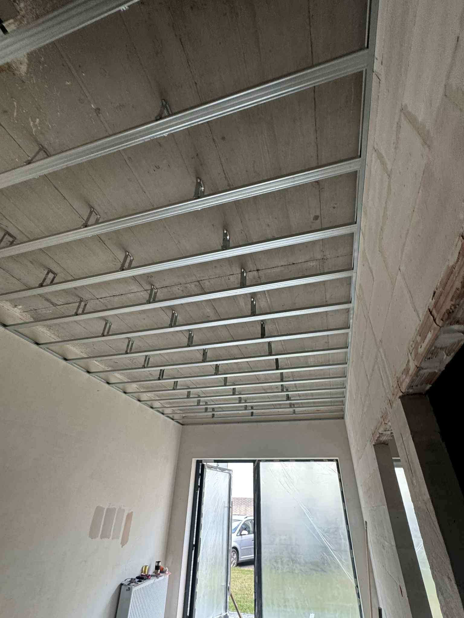 Szkielet sufitu podwieszanego z profili metalowych w surowym wnętrzu. Widoczne elementy mocujące, ściany w trakcie prac wykończeniowych, okno z folią ochronną.