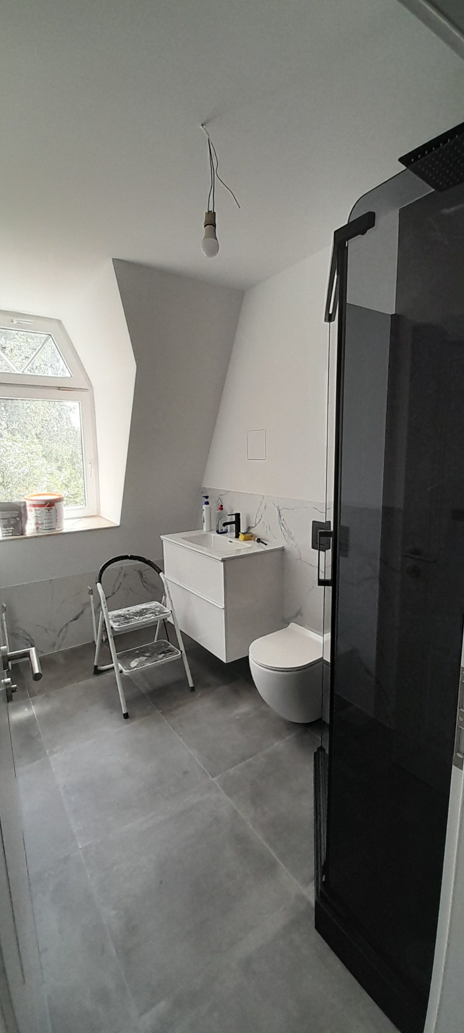 Wykończona łazienka na poddaszu z szarą podłogą, białą armaturą i kabiną prysznicową z czarnymi elementami. Widoczne okno dachowe i drabinka malarska.