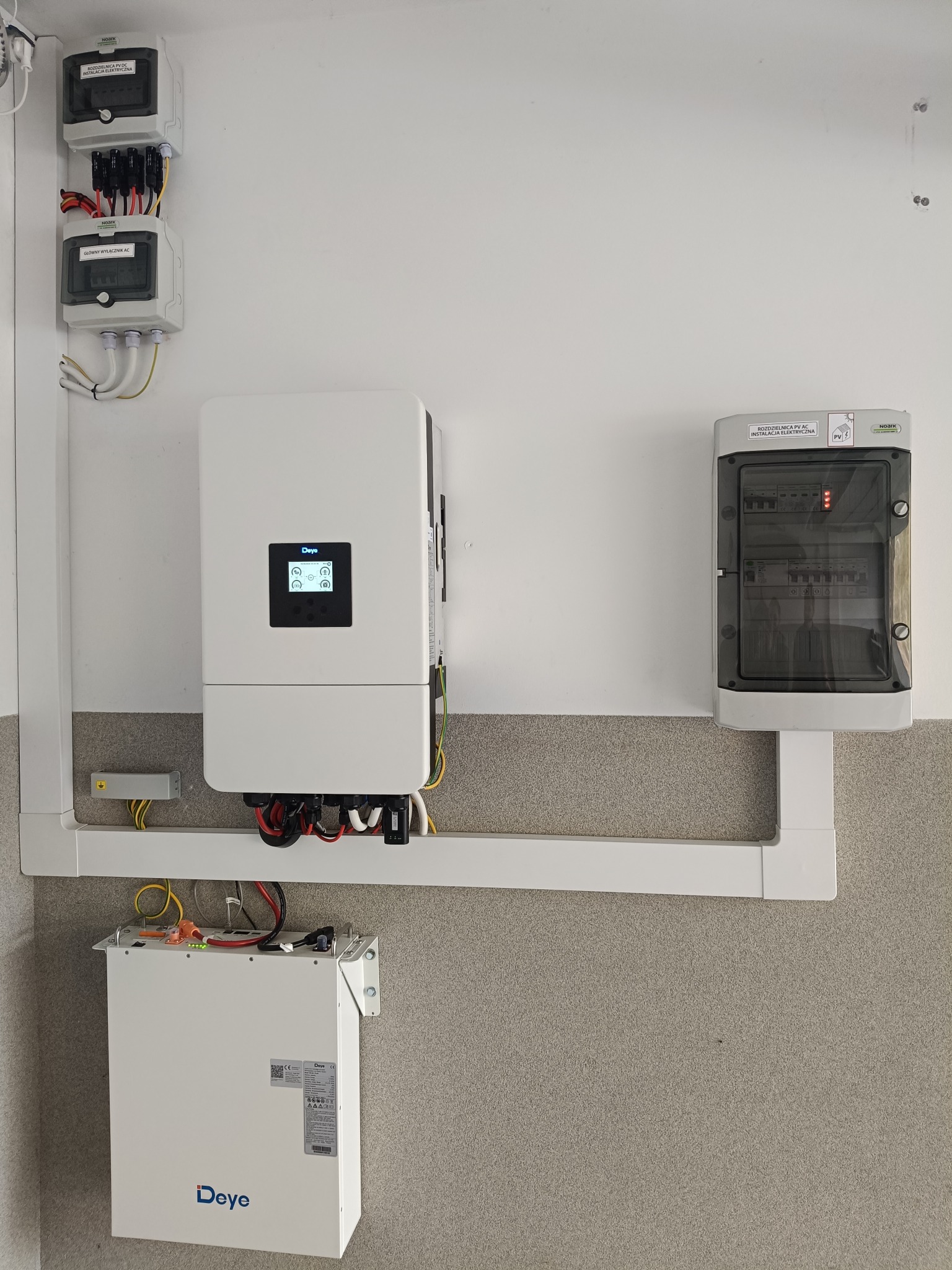 Instalacja: falownik Deye, magazyn energii i rozdzielnica obwodów gwarantowanych na ścianie. Widoczne kable i przewody. Szara ściana w tle.