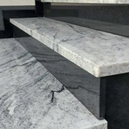 Granitowe schody zewnętrzne z szarego kamienia z wyraźnym wzorem żyłkowania, osadzone na czarnych, gładkich elementach. Widok z dołu, podkreślający strukturę kamienia.