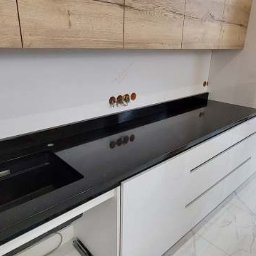 Bednarz & Stone Tomasz Bednarz - Czarny, błyszczący blat kamienny z wbudowanym zlewem w nowoczesnej kuchni z białymi szafkami i drewnianymi elementami. Widoczne otwory na baterię i akcesoria.