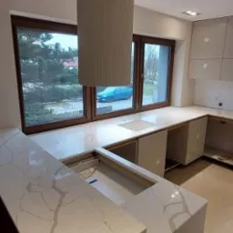 Jasny blat kuchenny z subtelnym wzorem marmuru, z otworem na zlew, pod oknem. Nowoczesny okap cylindryczny. Minimalistyczna kuchnia w neutralnych barwach.