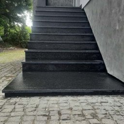 Bednarz & Stone Tomasz Bednarz - Ciemne, kamienne schody zewnętrzne prowadzące do budynku z szarą elewacją. Stopnie o gładkiej powierzchni, ułożone na zewnątrz na kostce brukowej. Minimalistyczny design.