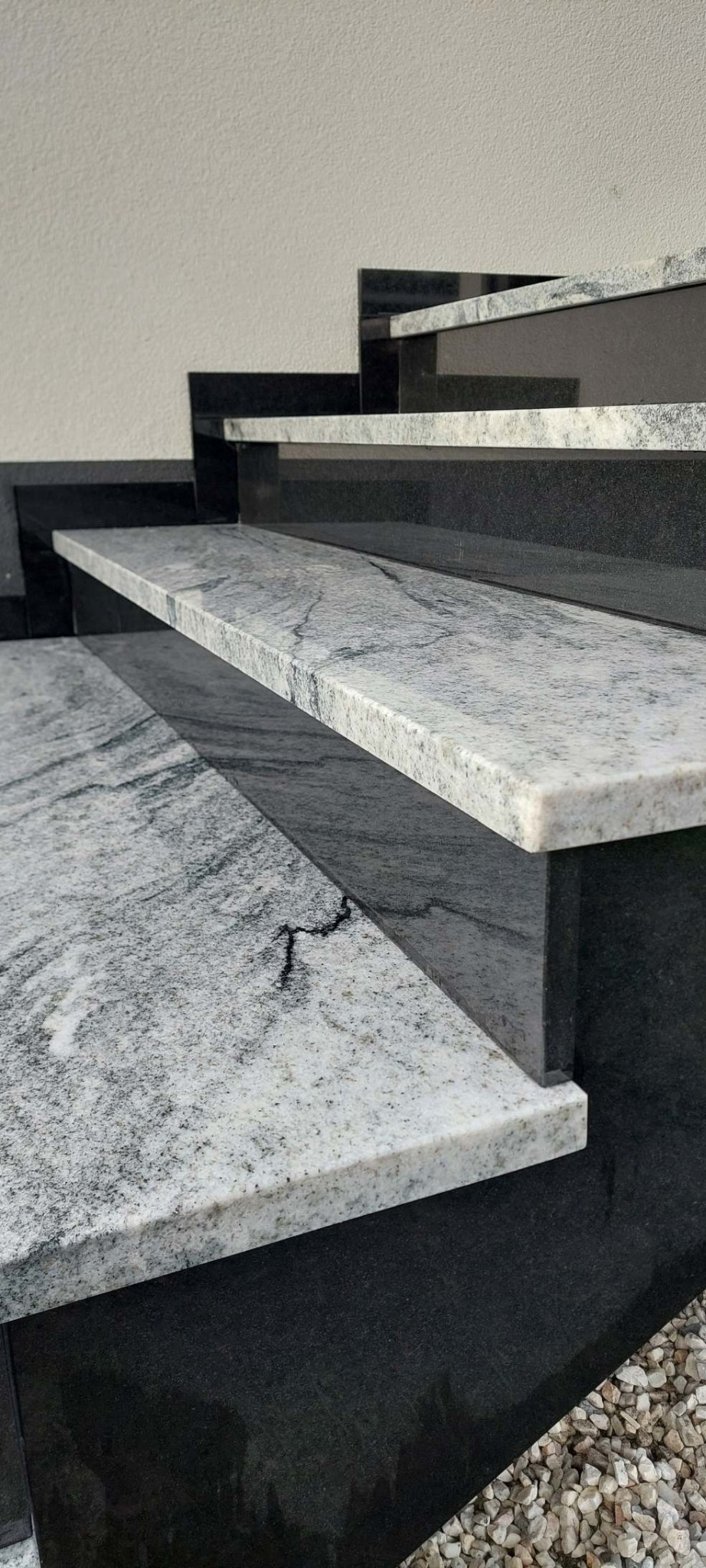Granitowe schody zewnętrzne z szarego kamienia z wyraźnym wzorem żyłkowania, osadzone na czarnych, gładkich elementach. Widok z dołu, podkreślający strukturę kamienia.