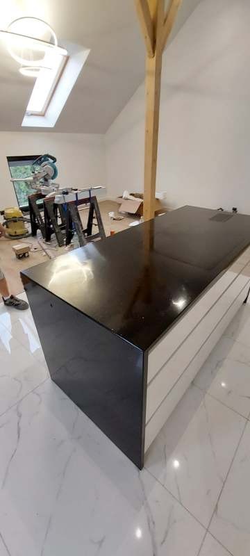 Błyszczący, czarny blat kuchenny z widocznym fragmentem białych szafek w jasnym wnętrzu z marmurową podłogą i narzędziami w tle. Ukończony montaż.