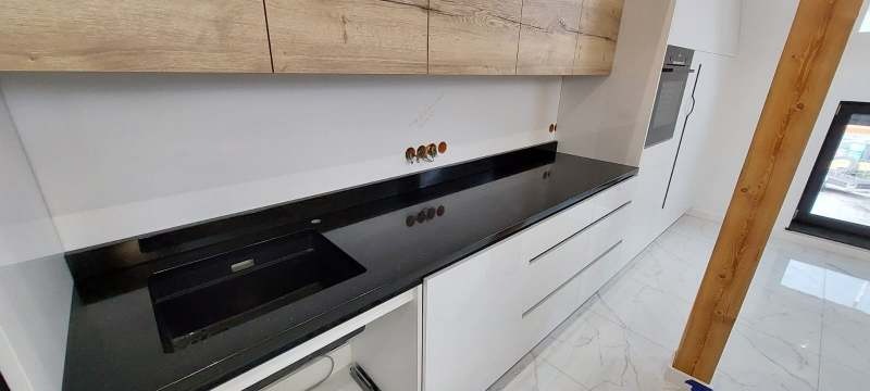Czarny, błyszczący blat kamienny z wbudowanym zlewem w nowoczesnej kuchni z białymi szafkami i drewnianymi elementami. Widoczne otwory na baterię i akcesoria.