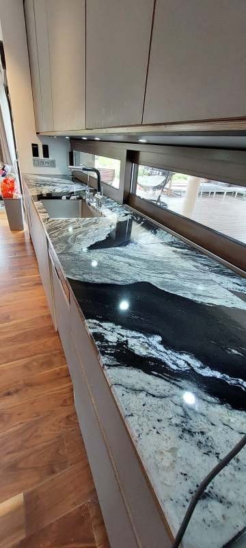 Czarno-biały blat kamienny z połyskiem, zamontowany w kuchni z oknem. Widoczny zlew i bateria. Blat o unikalnym wzorze przypominającym marmur.