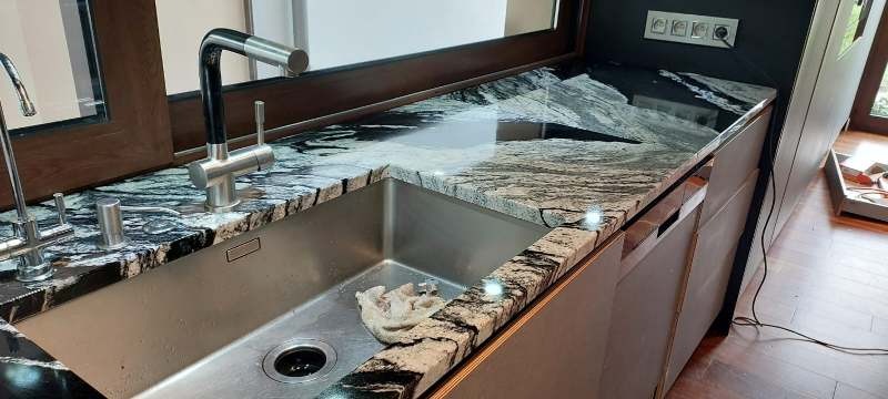 Czarno-biały blat kamienny z połyskiem, zintegrowany ze stalowym zlewem i nowoczesną armaturą. Widoczne gniazdka elektryczne w tle.