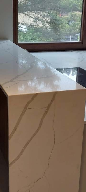 Błyszczący, beżowy blat kamienny z szarymi żyłkami, widoczny fragment płyty indukcyjnej i okno z widokiem na zieleń. Nowoczesny design, gładka powierzchnia.