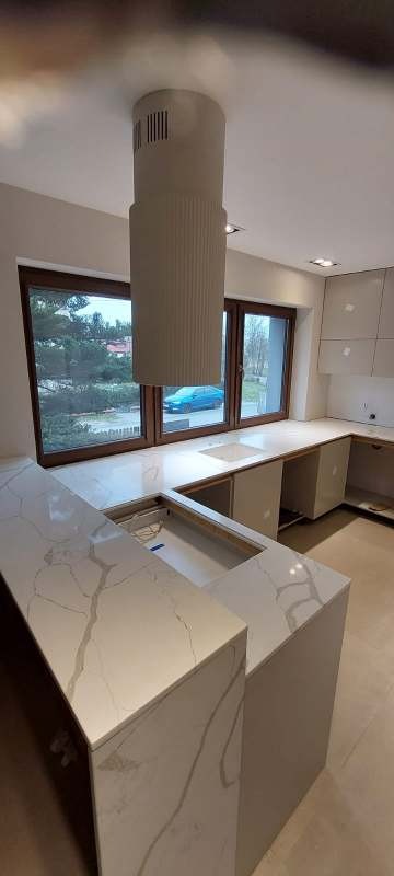 Jasny blat kuchenny z subtelnym wzorem marmuru, z otworem na zlew, pod oknem. Nowoczesny okap cylindryczny. Minimalistyczna kuchnia w neutralnych barwach.