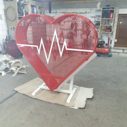 Usługi ślusarsko-spawalnicze Ciesiółka Jacek - Czerwona, metalowa konstrukcja w kształcie serca z białym motywem EKG, umieszczona na białym stelażu. Wykonana z siatki. Stoi w warsztacie na kartonie.