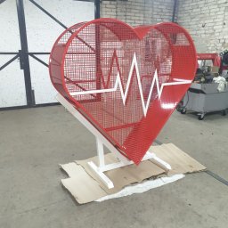 Usługi ślusarsko-spawalnicze Ciesiółka Jacek - Czerwona, metalowa konstrukcja w kształcie serca z białym symbolem EKG, umieszczona na białym stelażu w warsztacie. Nietypowa dekoracja.