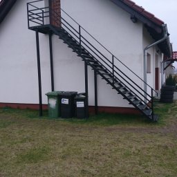 Usługi ślusarsko-spawalnicze Ciesiółka Jacek - Zewnętrzne schody metalowe z czarną, prostą balustradą prowadzące do drzwi na piętrze budynku. Na pierwszym planie widoczne pojemniki na śmieci i trawnik.