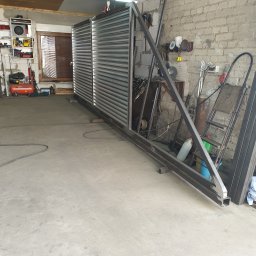 Usługi ślusarsko-spawalnicze Ciesiółka Jacek - Warsztatowa fotografia ukończonej, metalowej konstrukcji bramy z wypełnieniem z blachy trapezowej, oparta o ścianę z cegły. Widoczne narzędzia i akcesoria ślusarskie.