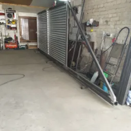 Warsztatowa fotografia ukończonej, metalowej konstrukcji bramy z wypełnieniem z blachy trapezowej, oparta o ścianę z cegły. Widoczne narzędzia i akcesoria ślusarskie.