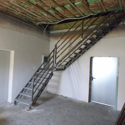 Usługi ślusarsko-spawalnicze Ciesiółka Jacek - Metalowe schody z ażurowymi stopniami w surowym wnętrzu. Widoczne ocieplenie stropu i narzędzia na podłodze. Ujęcie z dołu, podkreślające konstrukcję.