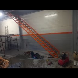 Usługi ślusarsko-spawalnicze Ciesiółka Jacek - Pomarańczowe schody metalowe przymocowane do ściany w hali. Widoczne narzędzia i kompresor. Hala z szarą podłogą i białą ścianą z pomarańczowym pasem.