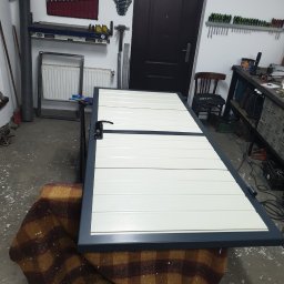 Usługi ślusarsko-spawalnicze Ciesiółka Jacek - Biała, segmentowa brama garażowa w trakcie montażu w warsztacie. Widoczne narzędzia i elementy konstrukcyjne. Brama oparta o koc w kratę.