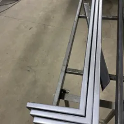 Kątowniki ze stali, trzy srebrne profile ułożone na metalowym stojaku w warsztacie. Widoczne ślady obróbki i surowy beton na podłodze.