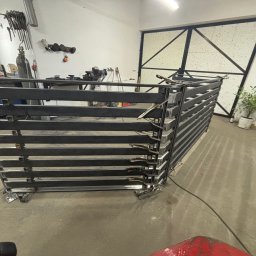 Usługi ślusarsko-spawalnicze Ciesiółka Jacek - Warsztatowe ujęcie metalowej konstrukcji z poziomych profili, zabezpieczonej klamrami spawalniczymi, na tle białej ściany z narzędziami i metalowej bramy.