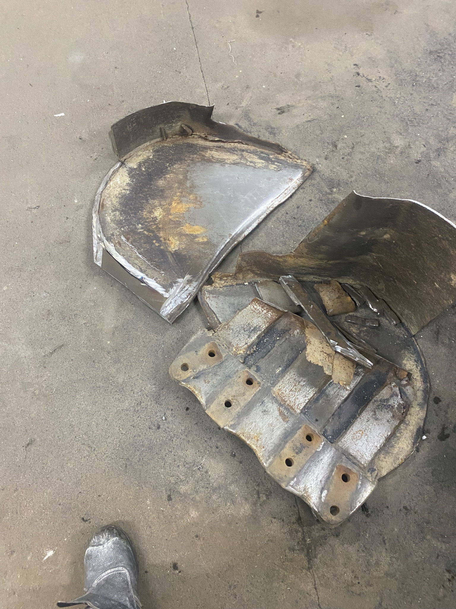 Uszkodzony, metalowy element maszyny budowlanej, prawdopodobnie łyżka koparki, leżący na betonowej posadzce. Widoczne ślady korozji i spawania. Fragment buta w lewym dolnym rogu.