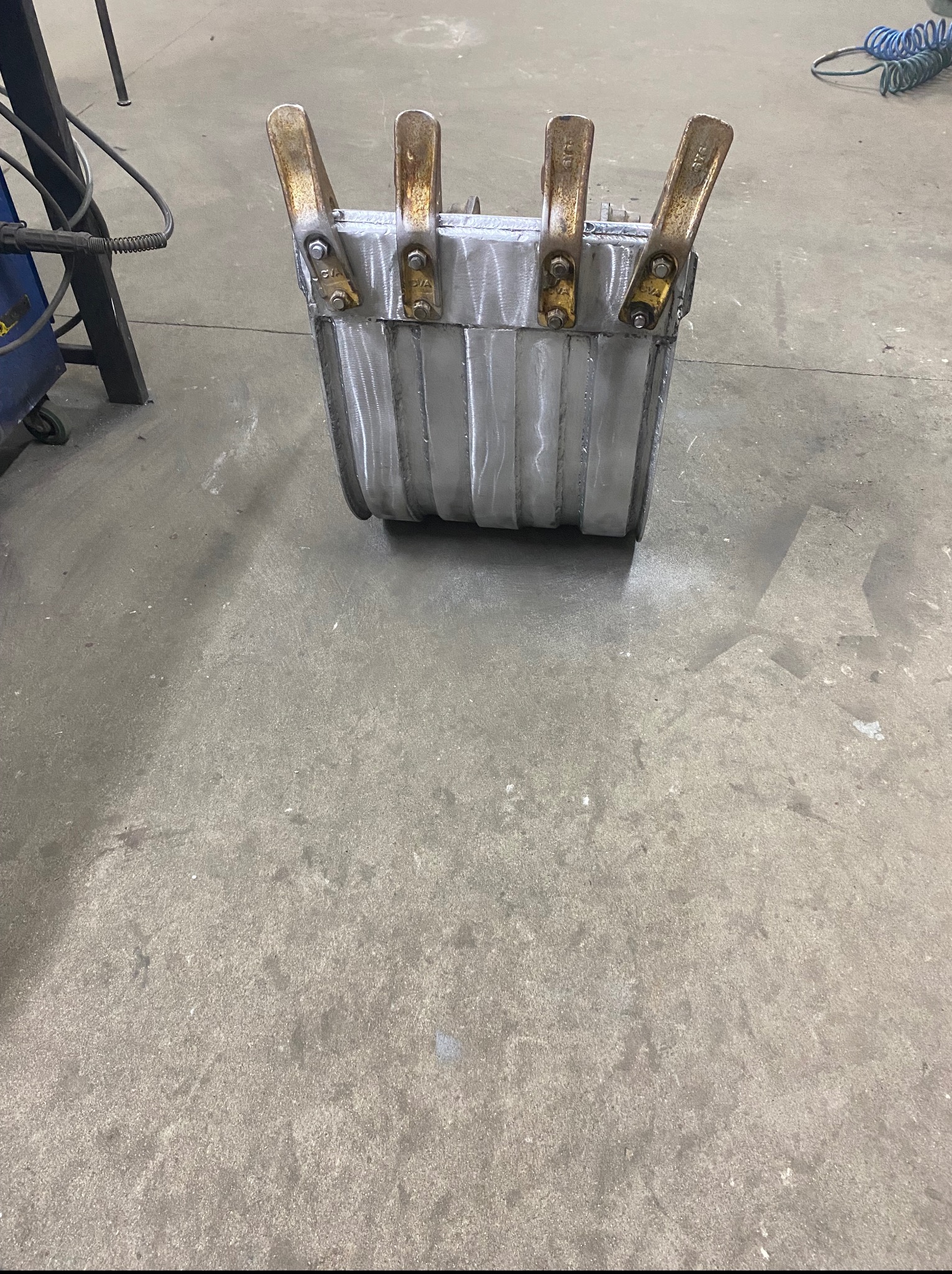 Metalowa konstrukcja z widocznymi spawami, zabezpieczona czterema zaciskami śrubowymi, stojąca na betonowej posadzce w warsztacie. Detal z bliska.